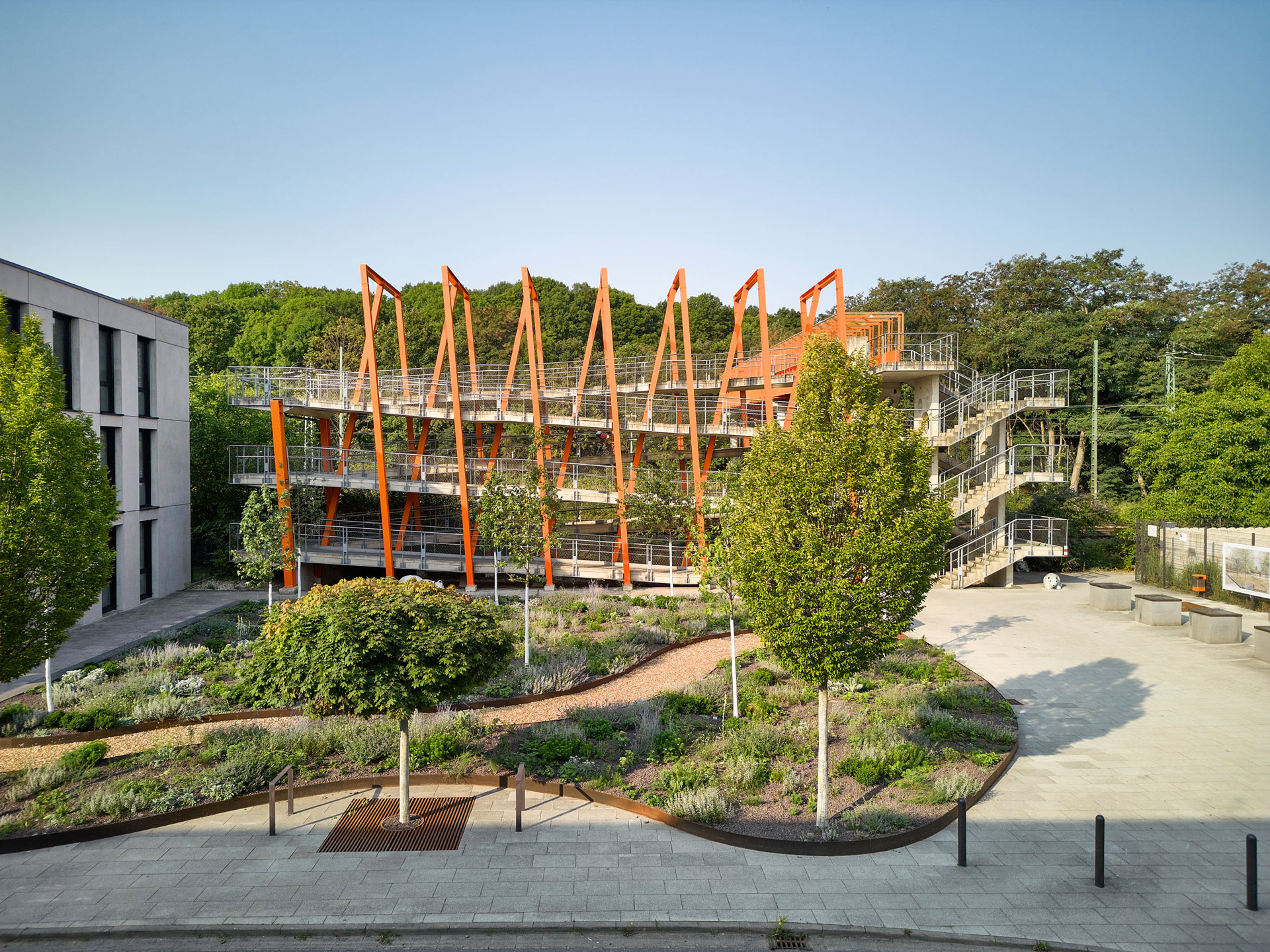 Der Belvedere Platz mit Bäumen und bepflanzten Beeten vor der Belvedere-Brücke mit architektonisch auffälligen orangefarbenen Stahlträgern bei blauem Himmel.